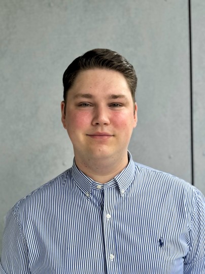 Auf dem Portraitfoto ist der Junior Sales Manager zu sehen. Ein braun haariger junger Mann mit einem hellblauen Hemd.